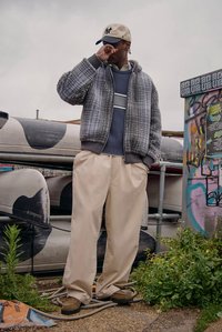 Šedá texturovaná bunda, modrý proužkovaný svetr, béžové kalhoty s širokými nohavicemi a hnědé boty. Osoba s světlou čepicí stojí u graffiti.