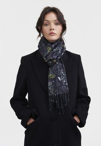 Sciarpa a stampa floreale scura realizzata in tessuto morbido, con colori vivaci su base blu navy, con estremità frangiate. Indossata con un cappotto nero.