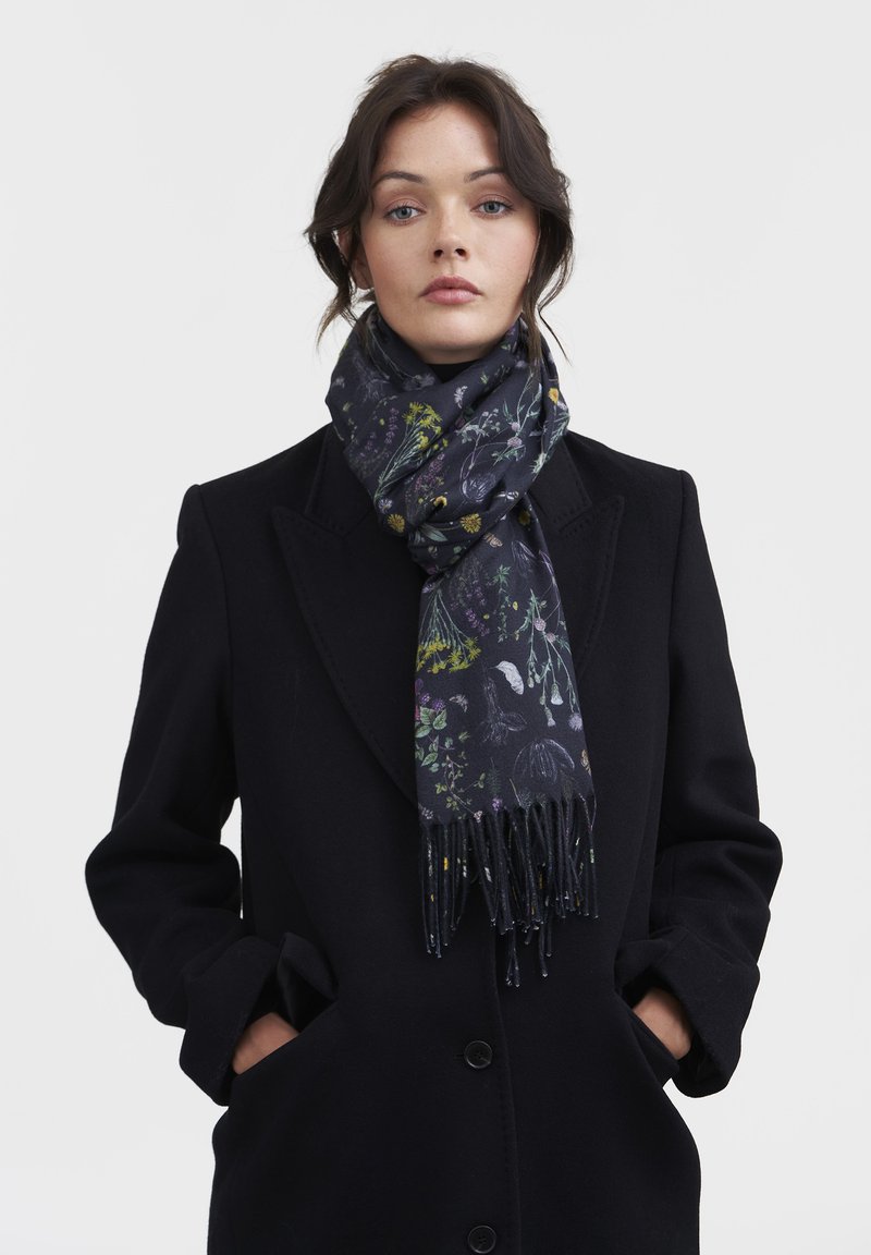 Sciarpa a stampa floreale scura realizzata in tessuto morbido, con colori vivaci su base blu navy, con estremità frangiate. Indossata con un cappotto nero.