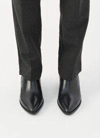 Μαύρες δερμάτινες μπότες μέχρι τον αστράγαλο με μυτερή μύτη, λείο υφή και ελαφριά γυαλάδα, συνδυασμένες με σκούρες γκρι κομψές παντελόνες.