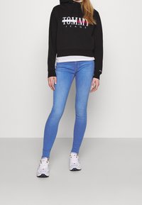 Sudadera recortada negra con el logo "TOMMY JEANS", combinada con vaqueros skinny de color azul claro y zapatillas blancas con detalles negros.