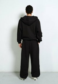 Mann mit lockigem Haar steht mit dem Gesicht zu einer weißen Wand, trägt einen lockeren schwarzen Hoodie und weite schwarze Hosen mit weißen Turnschuhen.