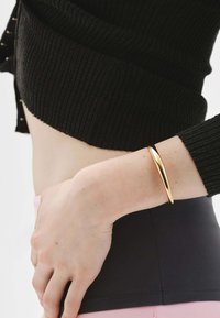 Brazalete de pulsera de oro con un acabado liso y pulido, llevado en una muñeca contra una blusa negra de canalé y pantalones oscuros ajustados.
