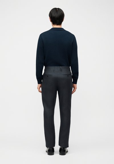 Άνδρας όρθιος στραμμένος προς τα πίσω, φορώντας μπλε ναυτικό πουλόβερ, σκούρο γκρι παντελόνια με πίσω τσέπες και μαύρα επίσημα παπούτσια, μπροστά σε απλό φόντο.