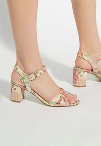 Floral-patterned block heel sandals with adjustable ankle strap, featuring light pastel colors and textured material. Accented with gold hardware.