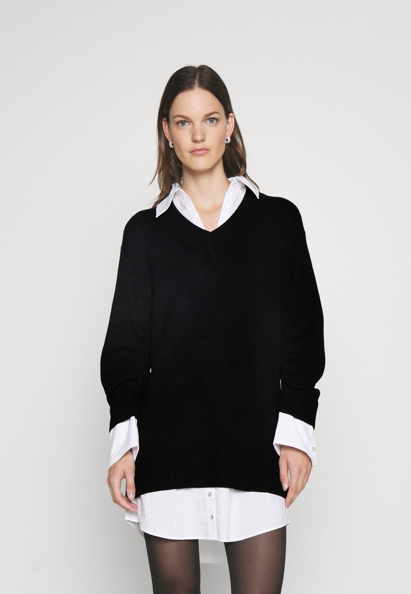 Schwarzer gestrickter Pullover mit V-Ausschnitt, gerippten Bündchen und Saum, über einem weißen Hemd getragen. Das Model trägt transparente schwarze Strumpfhosen.