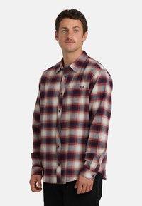 Camicia in flanella a quadri rossi e blu navy con maniche lunghe e chiusura a bottoni, dotata di una tasca sul petto e un orlo leggermente curvato.