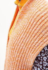 Pletená oranžová a broskvová vesta s texturovaným vzorem, spárovaná s hnědou košilí s květinovým potiskem, na které jsou bílé květy a akcenty.