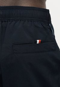 Κοντινό σε μαύρο σορτς με ελαστική μέση και πίσω τσέπη που έχει μια μικρή κόκκινη, λευκή και μπλε ριγέ ετικέτα.