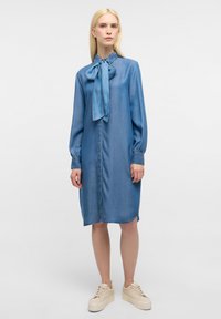 Abito di denim con maniche lunghe, dettaglio con fiocco al colletto e apertura frontale con bottoni. Colore: azzurro chiaro. Indossato con sneakers beige con piattaforma.