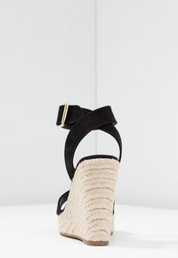 Sandalia de cuña negra de ante con un tacón de cuerda blanca texturizada, correa en el tobillo ajustable y detalle de hebilla dorada. Vista trasera que muestra el diseño curvado.