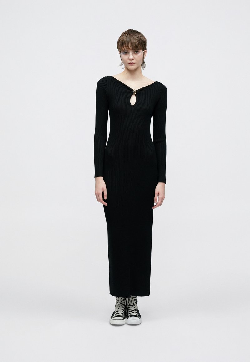 Rochie neagră din tricot cu model în dungi, croială potrivită, mâneci lungi, decupaj în formă de cheie la decolteu și lungime maxi dreaptă. Asortată cu adidași negri.