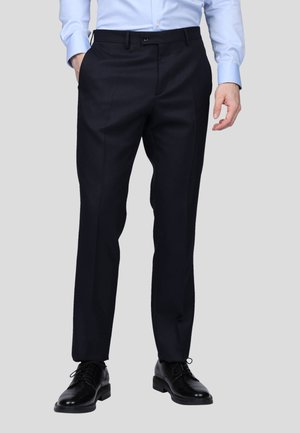 Pantalons de costume navy dans une coupe ajustée, avec poches latérales, un devant plat et une texture lisse. Portés avec des chaussures de dress noires brillantes.
