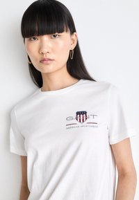 T-shirt bianco in cotone con colletto tondeggiante, caratterizzato da un logo ricamato multicolore che include un design a scudo e del testo. Maniche corte.
