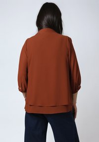 Vrouw met lang donker haar die een roestkleurige blouse met lange mouwen en donkerblauwe broek draagt, gezien van achteren tegen een effen achtergrond.