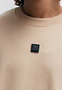 Sudadera beige con un cuello acanalado que presenta un parche negro cuadrado grabado con "HUGO" en letras plateadas. Textura suave, diseño casual.