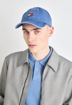 Young man wearing a blue Tommy Jeans cap, light blue polo shirt, and light gray zip-up jacket, looking directly at the camera.