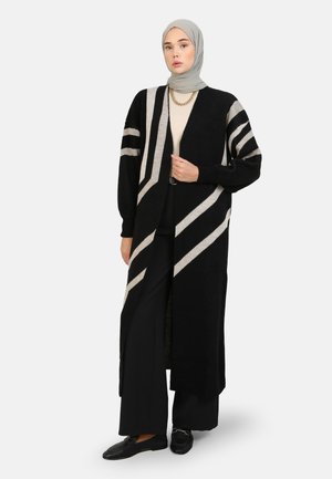 Cardigan long noir avec des rayures géométriques crèmes, fabriqué en tissu texturé. Il présente une coupe décontractée et un design ouvert à l'avant pour un superposition facile.