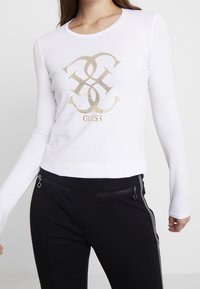 Camicia bianca a maniche lunghe con un design grafico dorato metallico e la parola "GUESS" centrata sotto. Accoppiata a pantaloni neri con tasche.