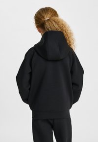Svart oversized hoodie med dragsko på huvan, med ribbad muddar och nederdel, tillverkad av mjukt tyg. Bärs med matchande svarta byxor.