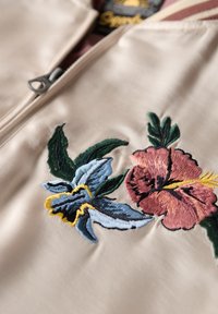 Kremowy materiał z zamkiem błyskawicznym, ozdobiony żywą haftowaną florą w odcieniach różu, niebieskiego i zielonego, z starannym szyciem oraz teksturowymi akcentami.
