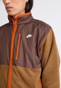 Nike jakna s gornjim dijelom od smeđeg, glatkog najlona s narančastim zatvaračem, naglašena donjim dijelom od flisa u svjetlijoj smeđoj boji. Džep na prsima s zatvaračem.
