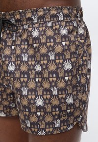 Καφέ, λευκά και μπεζ μαγιό με μοτίβο από τροπικά φύλλα και λουλούδια, με ελαστική μέση και μαύρο κορδόνι σε άτομο.