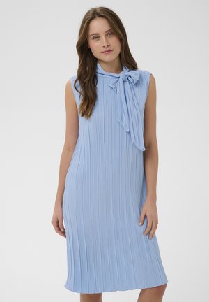 Kobieta w bezrękawnikowej, jasnoniebieskiej plisowanej sukience z dużą kokardą zawiązaną na szyi, stojąca na tle jasnego, jednolitego tła.
