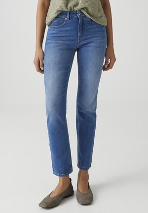 Vrouw draagt lichtblauwe skinny jeans, olijfgroen gebreid topje en grijze platte schoenen, staand op een effen lichte achtergrond.