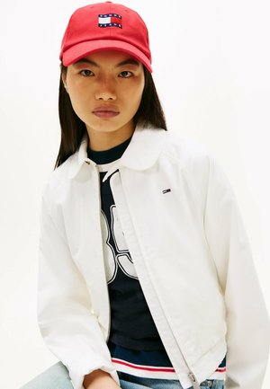 Giovane con cappellino rosso Tommy Jeans, giacca bianca con zip sopra una t-shirt nera con grafica e jeans azzurri, posando davanti a uno sfondo bianco.