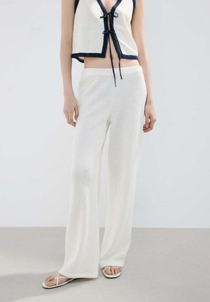 Kobieta ubrana w białe, teksturowane spodnie o szerokich nogawkach, dopasowany bezrękawnik crop top z granatowym obszyciem i troczkami oraz białe klapki.