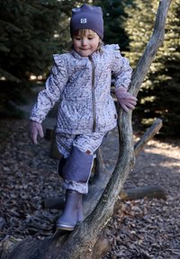 Lila Wintermantel mit Blumenmuster und Rüschendetails, passende Hose, Handschuhe und graue wasserdichte Stiefel, getragen von einem Kind auf einem Baumstamm.