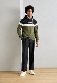 Olivengrønn og svart hettegenser med hvit stripe-design, kombinert med svarte bukser og hvite sneakers, med snor i hetta.