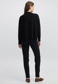 Schwarzer oversized Pullover mit einer weichen Textur, der überschnittene Schultern und seitliche Schlitze aufweist. Kombiniert mit schwarzen Slim-Fit-Hosen und Schuhen mit Tierdruck.