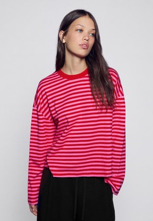 Jeune femme aux longs cheveux bruns portant un t-shirt à manches longues rayé rouge et rose et un pantalon noir sur un fond uni.