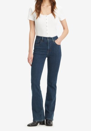 Frau trägt weißes kurzärmeliges, geknöpftes Oberteil, blaue hoch taillierte Jeans mit Nadelstreifen und schwarze Stiefeletten mit Absatz, steht mit einer Hand in der Tasche.