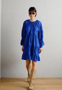 Blå lager-på-lager klänning med långa, puffiga ärmar och en rund halsringning. Modellen bär solglasögon och knähöga beige stövlar på ett trägolv.