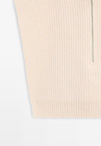 Krämfärgad stickad tyg med ribbad nederkant och en vertikal silverdragkedja på högra sidan.