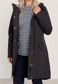 Czarna parka z zamkiem błyskawicznym i zapięciem na napy z futrzanym kapturem, dwiema kieszeniami bocznymi i średniej długości krojem, noszona na wzorzystej niebieskiej bluzce.