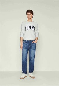 Jovem vestido com um suéter branco "TOMMY", jeans azuis e ténis brancos, encostado a um fundo claro e liso, com uma mão no bolso.