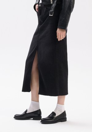 Black denim skirt with a front slit, frayed hem, and pockets, paired with black loafers and grey socks. Tailored fit and mid-length design.