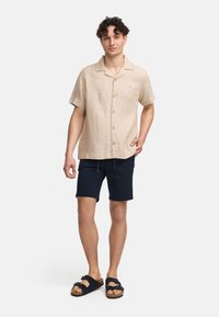 Camisa corta de lino color tostado con botones, combinada con pantalones cortos azul marino y sandalias negras. La camisa tiene un corte relajado y un cuello plano.