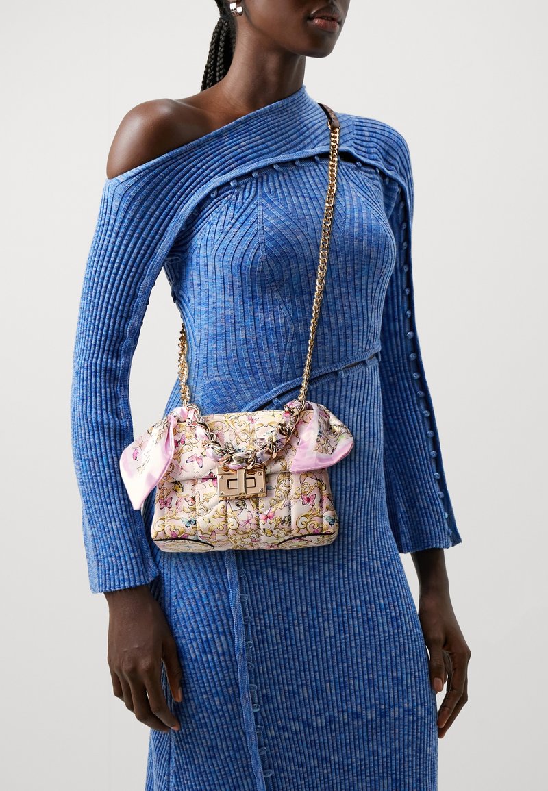 Femme portant une robe bleue côtelée à épaules dénudées, portant un sac matelassé fleuri avec une chaîne dorée et un foulard rose noué de chaque côté.
