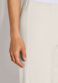 Linkerhand ontspannen naast de heup, in hooggerande crèmekleurige broek met verticale naaddetails, neutrale achtergrond.