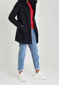 Persona con un cappotto nero con cappuccio, maglione rosso, jeans arrotolati di un blu chiaro e sneakers bianche, in piedi con le mani nelle tasche del cappotto.
