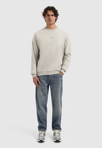 Jonge man met krullend haar, gekleed in een beige sweatshirt met de tekst "PURE PATH", blauwe spijkerbroek en grijze sneakers, staand tegen een effen achtergrond.