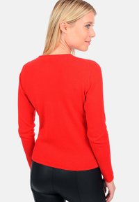 Roter Pullover mit langen Ärmeln, Rundhalsausschnitt, glatter Textur, taillierter Passform und Schulternahtdetails. Unterhalb sichtbar sind schwarze Hosen.