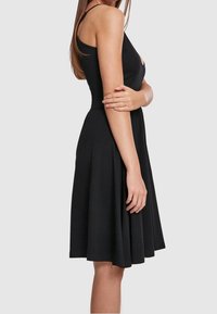 Svart ärmlös klänning med en åtsittande bodice och utställd kjol, gjord av mjukt tyg. Har en blygsam halsringning och naturlig drapering.