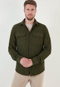 Cămașă de un verde închis, cu revere, cu buzunare cu nasturi pe piept, hem rotunjit și mâneci lungi, purtată peste un tricou de o culoare deschisă.