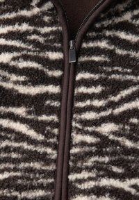 Tissu moelleux marron et crème avec une fermeture éclair métallique à l'avant et des finitions marron foncé, ressemblant à un motif imprimé animal texturé.